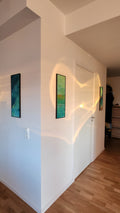 Two narrow vertical paintings with turquoise tones and gold leaf installed in a minimalist Scandinavian hallway, with warm sunlight reflections on white walls and wooden floor