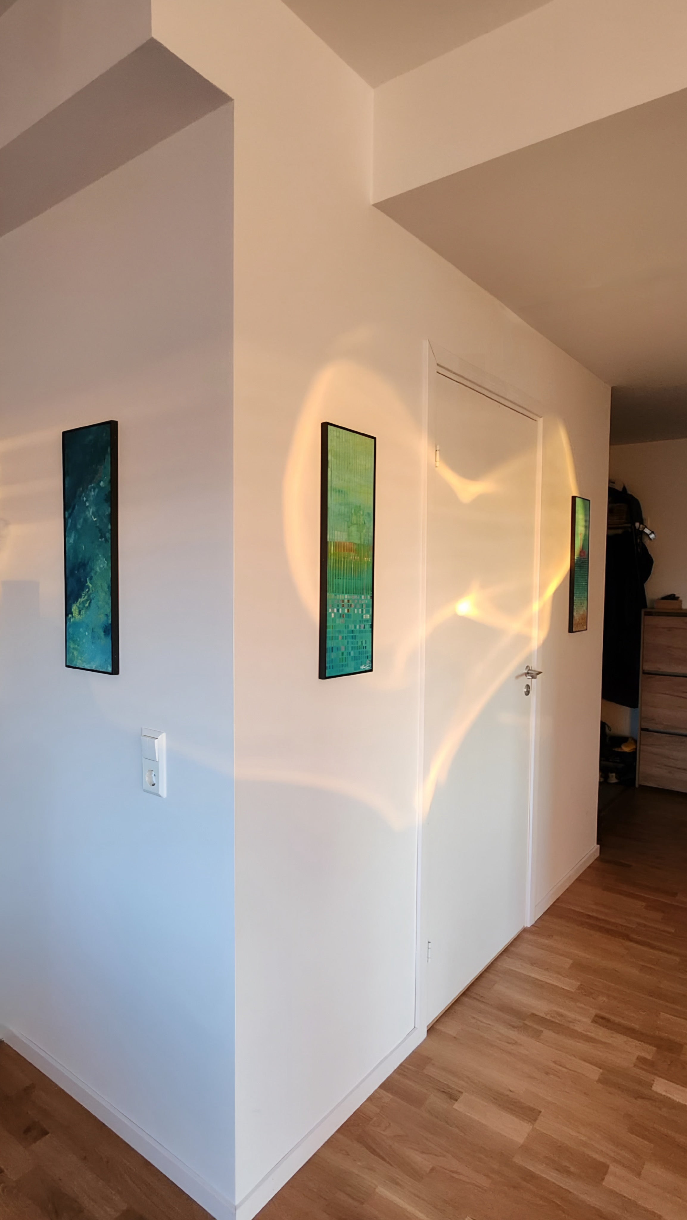 Two narrow vertical paintings with turquoise tones and gold leaf installed in a minimalist Scandinavian hallway, with warm sunlight reflections on white walls and wooden floor
