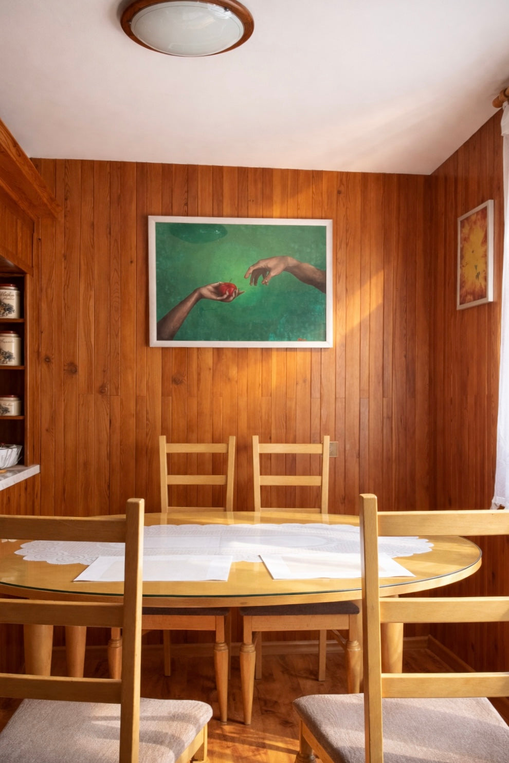 Tempera painting of two hands with a red apple, installed above a wooden dining table in a warm, sunlit interior with wooden wall panels.