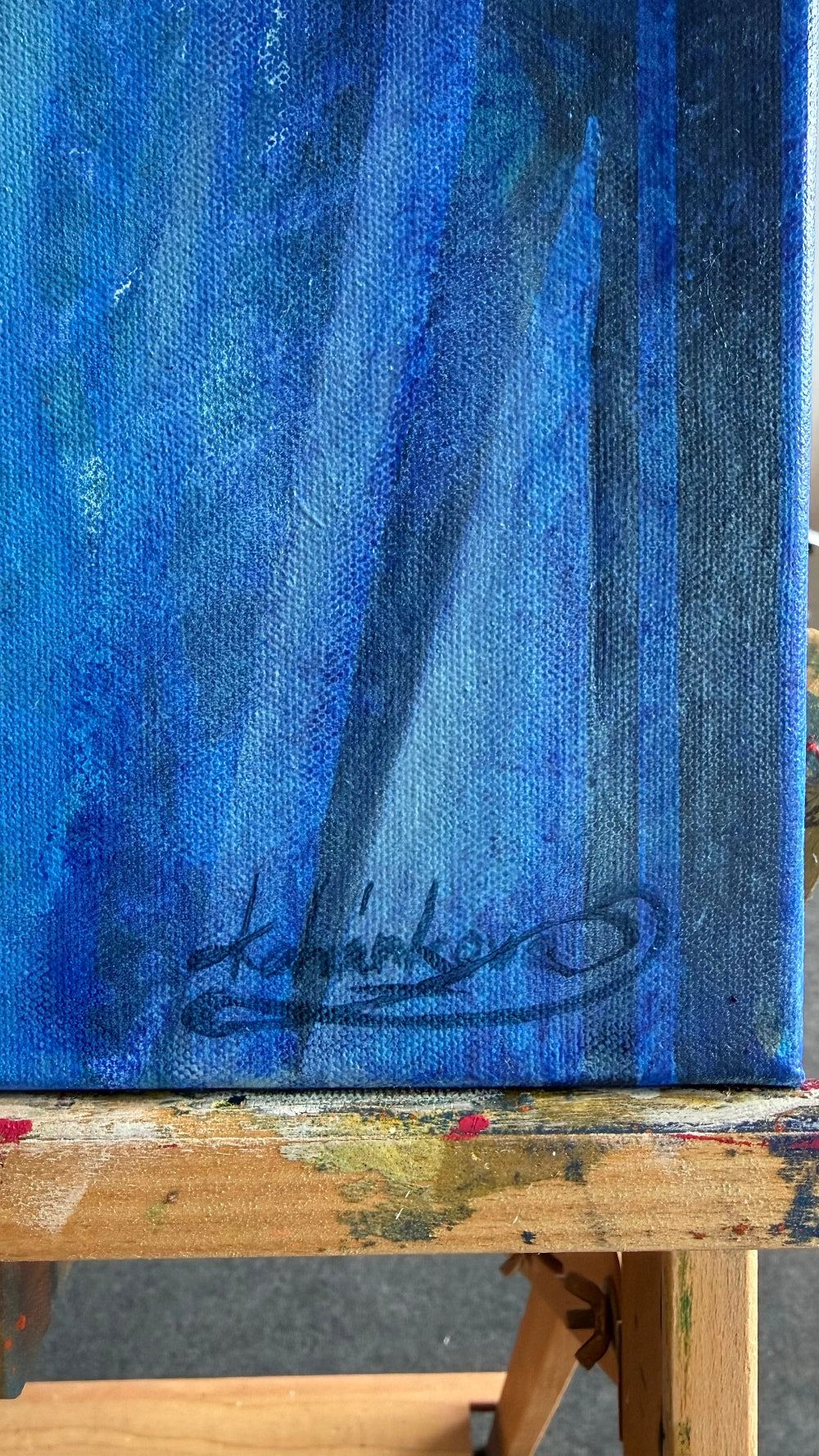 A close-up of a blue textured canvas painting on an easel, with a black artist signature near the bottom and the wooden easel visible around the edges.