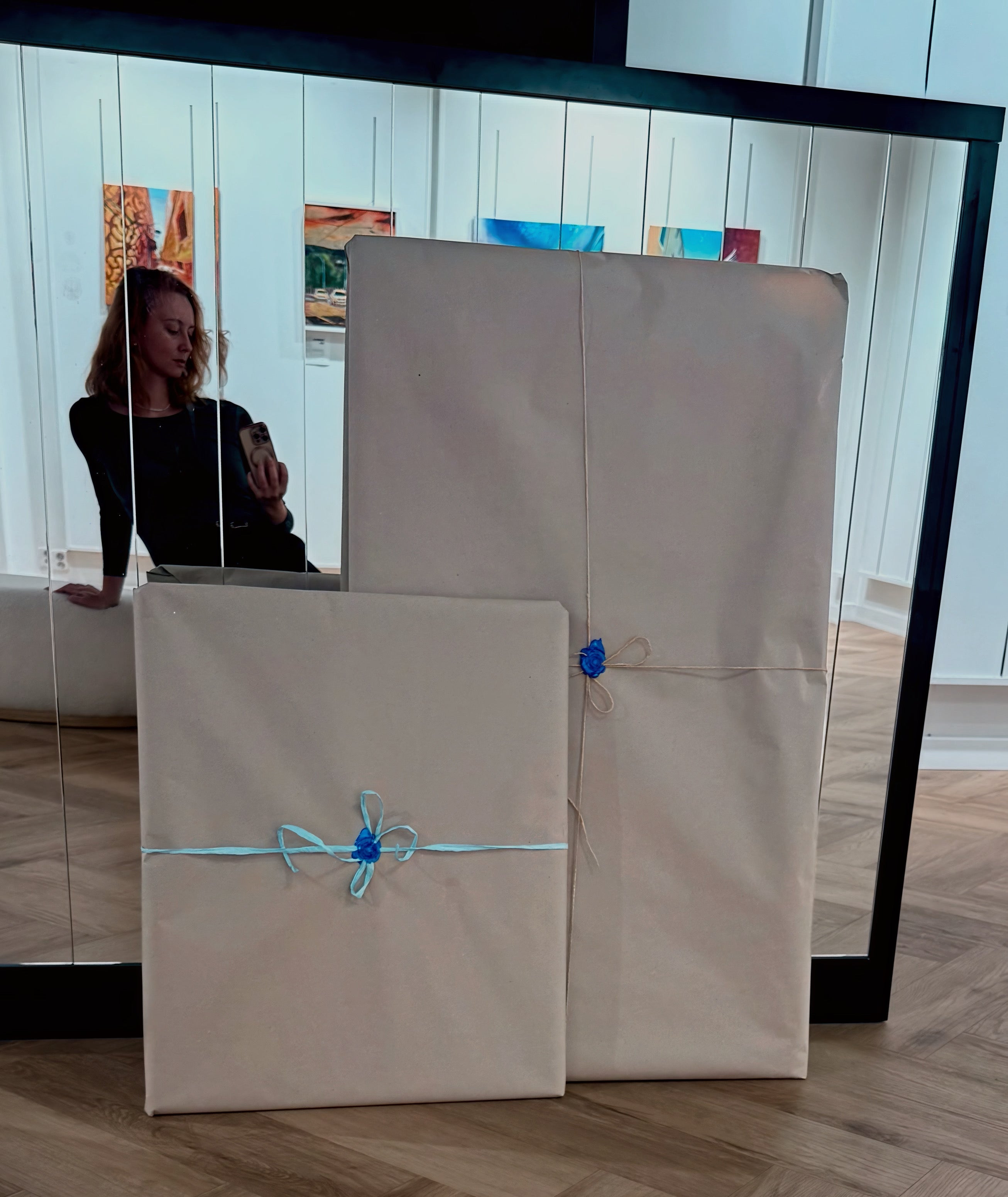 Two wrapped painting in packages on a wooden floor with a mirror reflecting a artist and artwork.